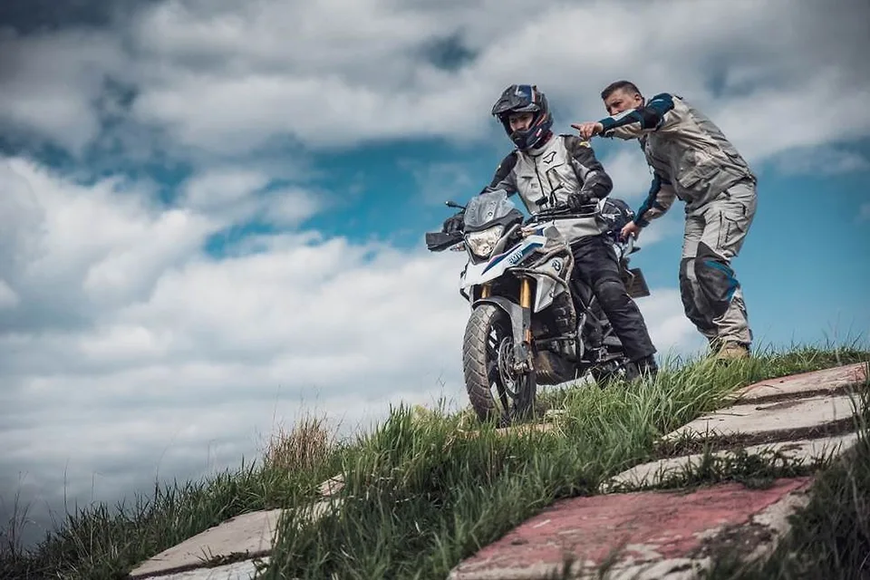Instruktor pomáhá jezdci na BMW GS zvládnout výjezd do svahu pod dramatickou oblohou
