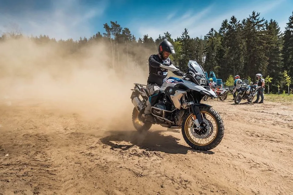 Jezdec na BMW GS zvedá písek na lesní terénní ploše, v pozadí stojící jezdci